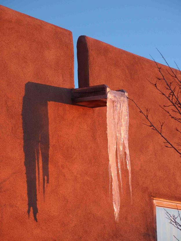 {Image:  a flat water spout protrudes from an adobe wall with several feet of frozen water suspended off the end.  The adobe is much darker than the previous photo because of the setting sun.}}