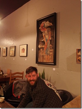 {Image:  photo of a white man with short brown hair and a beard sitting in an upholstered chair looking toward the camera.  Above and to his right is a painting perhaps 3 feet high by 2 feet wide of a person standing facing the viewer with his hands held out in front of him as if to show something.  In the background of this large painting are buildings.  In the background of the photo are additional, smaller paintings and other chairs and tables in a coffee shop.}