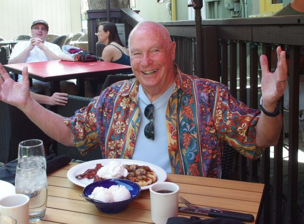 Image:  older white man in a flowered shirt sitting at an outdoor restaurant table, smiling with his arms outstretched.  On the table in front of him is a plate of waffles and bacon, with ice cream on top.
