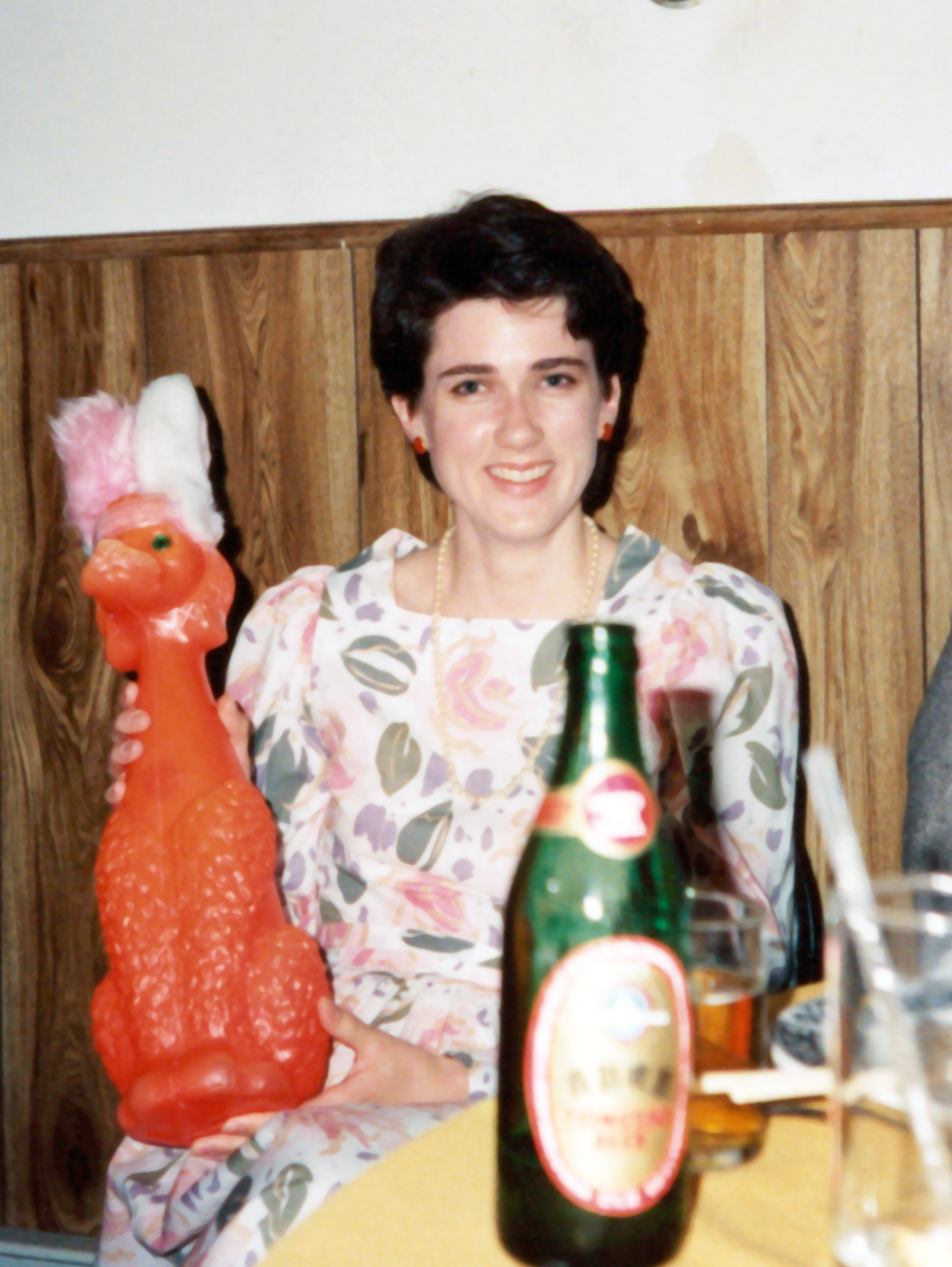 Image: Photo of me at age 28, white woman with short dark hair wearing a flowered dress, holding an orange plastic poodle with pink fuzzy rabbit ears. I'm sitting at a table with a bottle of beer in the foreground.